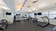 This single-story fitness facility is thoughtfully designed to accommodate a wide range of wellness activities across several distinct zones. The main cardio area is bright and spacious, featuring multiple treadmills, elliptical machines, and stair climbers arranged beneath vaulted ceilings fitted with ceiling fans and linear fluorescent lighting. A full-length wall mirror amplifies the sense of space, while multiple windows invite natural light throughout. Wall-mounted televisions provide entertainment during workouts.

A dedicated functional training room offers a more intensive experience, equipped with a cable machine, a large tire, agility ladder flooring, dumbbell racks, and industrial wall fans. This room features crown molding, generous window coverage, and rubber flooring.

A quiet lounge area with light wood flooring, soft gray walls, contemporary abstract artwork, and plush seating provides a comfortable space to rest. An adjoining meeting room includes a wall-mounted TV, an oval table, and bold blue chairs.

Additional amenities include a tanning room and a hallway hydration station with dual water fountains, a decorative wood-framed mirror, and wainscoting, connecting the various zones of this well-appointed facility.