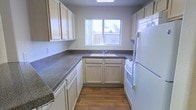 This single-floor kitchen is designed with a functional U-shaped layout that maximizes the use of available space. Light-toned wood cabinetry lines the walls both above and below the countertops, offering generous storage throughout. The dark, granite-style countertops provide a contrasting visual anchor and ample prep surface area. White appliances, including a refrigerator and range, complement the cabinetry and contribute to a clean, cohesive aesthetic. A stainless steel sink is positioned beneath a centered window, which allows natural light to fill the space and offers a view of the surrounding exterior. An overhead skylight further enhances the brightness of the room, creating a well-lit environment. The flooring consists of warm-toned hardwood-style planks that add a touch of warmth to the otherwise neutral palette. The overall ambiance is practical and tidy, with a straightforward design that prioritizes utility. The layout allows for efficient movement between the cooking, prep, and cleaning zones, making it a well-organized and functional kitchen space.