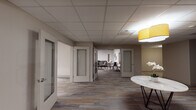 Located at 1 E Main St in Bay Shore, NY, this single-floor amenity space offers a thoughtfully designed environment that blends social and recreational functionality with modern aesthetics. The layout is organized around a central reception-style foyer, anchored by a striking marble-top round table beneath warm yellow pendant lighting and finished with multi-toned wood-look flooring that extends throughout the space.

A wide, well-lit corridor connects the foyer to multiple private rooms featuring frosted glass panel doors, providing versatile enclosed spaces suited for meetings or focused use. The hallway is accented with wall sconces and a clean, neutral color palette that creates a calm, professional atmosphere.

The open lounge and recreation area is a standout feature, boasting soaring ceilings with an exposed dark-painted industrial finish contrasted by track lighting and a contemporary geometric chandelier. This expansive area includes comfortable seating with a plush sofa, mid-century modern dining tables and chairs, a large framed wall-mounted Scrabble board as a decorative focal point, and curated artwork throughout. A full-size pool table with a vibrant blue felt surface anchors the game room portion of this space.

Together, the areas create a dynamic, inviting environment that is equally suited for relaxation, socializing, and recreation.
