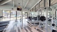 Located at 1501 E Grand Ave in Escondido, California, this fitness center is housed in a single-floor facility that makes a strong architectural impression from the moment you enter. The space is defined by soaring vaulted ceilings with exposed white-painted wood beam detailing and a white shiplap ceiling, creating an open, airy atmosphere that feels both expansive and inviting. Skylights and large floor-to-ceiling windows flood the interior with natural light, while ceiling fans and track lighting ensure comfort and visibility throughout.

The flooring features a stylish multi-tone wood-look plank pattern that adds a modern, clean aesthetic to the space. Full-length mirrors line the walls, amplifying the sense of space and allowing members to monitor their form during workouts.

The strength training area is comprehensively outfitted with a full rack of dumbbells, weight benches, squat racks, cable machines, and functional training equipment including TRX suspension trainers and medicine balls. Motivational fitness-themed typography art decorates the walls, adding an energetic visual element.

A dedicated cardio zone features modern treadmills and elliptical machines positioned directly in front of expansive windows, offering pleasant outdoor views of the surrounding landscape and pool area during workouts. The result is a thoughtfully designed, high-quality fitness facility built for performance and comfort.