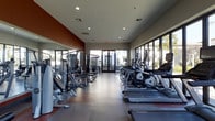 This fitness center is a thoughtfully designed, single-floor space that offers a comprehensive workout environment. The layout is generous, providing ample room between equipment stations to ensure comfortable movement throughout the facility. Large floor-to-ceiling windows line one side of the room, flooding the interior with natural light and offering views of the surrounding exterior, creating an open and energizing atmosphere.

On one side of the space, a dedicated strength training zone features a variety of cable machines and multi-use weight equipment, complemented by a full-length mirror wall that enhances the sense of depth and allows users to monitor their form. The opposite side is lined with a row of cardio machines, including ellipticals and treadmills, all oriented toward the windows to take advantage of the natural light and outdoor views.

The flooring transitions between rubberized mat sections near the weight equipment and a smooth, durable surface throughout the rest of the room. Recessed lighting in the ceiling supplements the abundant natural light. A bold accent wall in deep red adds a dynamic visual contrast to the otherwise neutral gray and white tones, contributing to a modern and motivating ambiance.