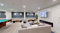 Located at Halifax St in Burnaby, BC, this well-appointed games room is a thoughtfully designed amenity space that balances recreation and relaxation. The open-plan layout accommodates several distinct zones, all unified by a neutral palette of crisp white walls, soft grey carpeting, and accents of slate blue that lend the space a modern, polished character.

At the heart of the room sits a full-sized pool table with a vivid green felt surface, illuminated by elegant brass pendant lights. Adjacent to it, a casual dining area features a round marble-top table surrounded by curved blue accent chairs, providing a social hub for gathering. A wood-framed cue rack and open shelving add functional detail to this zone.

The lounge area is anchored by a generous sectional sofa in light grey upholstery, positioned facing a wall-mounted flat-screen TV above a sleek linear electric fireplace — creating a warm, inviting atmosphere for relaxed viewing. A second large-screen TV is mounted in a separate nook framed by a natural wood slat feature wall, offering another entertainment option.

Recessed lighting throughout ensures the space is well-lit, while decorative wall sconces on the blue panelled accent wall add warmth. Potted indoor plants punctuate the room with greenery, and a built-in bench with wood top provides additional casual seating near the entrance.