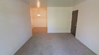 This single-floor space presents a straightforward, functional layout centered around open, connected living areas. The flooring throughout is covered in neutral-toned speckled carpet, providing a cohesive look across the rooms. White walls offer a clean, blank canvas, making the space easy to personalize or furnish to taste.

The main living area flows naturally into an adjacent room, partially visible in the background, where a pendant light fixture hangs from the ceiling, suggesting a designated dining area. This pendant light adds a subtle focal point and provides direct overhead illumination for that zone. A wall-mounted baseboard vent is visible, indicating a standard heating system.

A dark wood-toned door with a brass-finish handle is positioned along one wall, providing access to an additional room or exit point. The overall layout feels open and connected, with the transition between spaces marked by a subtle architectural division rather than full walls or doors.

The space maintains a simple, utilitarian aesthetic with minimal architectural detailing. The neutral palette and open flow make it highly adaptable, suitable for a variety of furniture arrangements and interior styles.