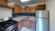 This single-floor unit offers a clean, functional layout with warm hardwood flooring throughout the living areas and bedrooms. The living room is bright and open, benefiting from multiple windows and a sliding glass door that leads directly to an outdoor deck, filling the space with natural light and providing easy access to the outdoors. A wall-mounted air conditioning unit and radiator heating ensure year-round comfort.

The kitchen is well-equipped and practical, featuring warm-toned wooden cabinetry that extends to the ceiling for ample storage, a mosaic tile backsplash, granite-style countertops, a stainless steel refrigerator, gas range, and a double sink. The flooring transitions to classic black-and-white tile, adding a timeless accent to the space.

The bedrooms are calm and understated, each with polished hardwood floors, windows offering views of surrounding greenery and neighboring structures, and individual radiator units for climate control. One bedroom also includes a wall-mounted air conditioning unit for added comfort.

The bathroom is compact and tidy, outfitted with a mirrored medicine cabinet featuring built-in shelving and overhead lighting, a wall-mounted toilet, and a sink with chrome fixtures. The neutral color palette throughout the unit creates a cohesive, clean aesthetic.