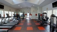 This fitness center offers a spacious and functional workout environment designed to accommodate a variety of exercise routines. The room is anchored by striking red and black checkered rubber flooring, providing both durability and a bold visual aesthetic underfoot. Vaulted ceilings rise overhead, lending the space an open, airy feel and enhancing the sense of volume throughout the room. Recessed lighting and natural light filtering in through windows keep the interior bright and welcoming. The cardio section is well-stocked with treadmills and elliptical machines positioned along one side, while a large wall-mounted mirror reflects the space and supports form-checking during workouts. On the opposite side, a cable-based strength training machine offers versatile resistance exercise options. White walls accented with red horizontal banding tie the color scheme together, reinforcing an energetic atmosphere. An emergency exit door is clearly marked at the rear of the room, ensuring safety compliance. The layout allows for comfortable movement between equipment, making the space accessible and practical for a range of fitness levels. Overall, this fitness center combines functional design with a motivating aesthetic to create an inviting space for daily exercise.