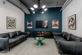 Located at 211 W Armour Blvd in Kansas City, Missouri, this thoughtfully designed single-floor amenity space blends industrial character with contemporary interiors. Exposed ceiling infrastructure, including visible ductwork and conduit, contrasts with refined finishes below, lending the space an urban, loft-inspired aesthetic throughout.

The central feature is a generously sized conference area anchored by a long natural wood table surrounded by black chairs, illuminated by a series of modern globe pendant lights. Curated artwork lines the soft blue-grey walls, adding a creative and professional atmosphere. Adjacent to the conference table, a warm café-style lounge nook is accessed through a curved arched opening, where teal banquette seating, cognac leather chairs, and round pedestal tables create an intimate setting ideal for informal gatherings.

A separate lounge area offers a more relaxed retreat, furnished with a sleek black leather sofa, a round green accent table, and a wall-mounted television set against a deep teal feature wall. Framed artwork and a mid-century media console complete the curated look of this zone.

Throughout the space, carpeted flooring in complementary tones delineates each functional area, while a glass-partitioned enclosure provides an additional private space. The overall design balances productivity and comfort, making it a versatile destination for collaboration, meetings, and social interaction.
