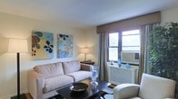 This single-floor residence offers a comfortable and thoughtfully designed living environment with a clean, contemporary aesthetic. The living room serves as the heart of the space, showcasing a neutral color palette anchored by soft, cream-toned upholstered seating arranged around a dark coffee table. Bold, colorful abstract artwork adorns the walls, adding vibrant visual interest and personality to the room. A window unit provides climate control, while a draped window with warm curtains allows natural light to filter into the space, creating a bright and welcoming ambiance. A classic table lamp adds warm, ambient lighting during evening hours. Decorative accents, including glass vessels, ceramic vases in calming blue tones, and a sculptural bowl, are carefully placed throughout, lending the space a curated, stylish character. The layout is efficiently organized, making excellent use of the available floor space to balance functionality with comfort. The overall atmosphere is relaxed yet polished, making it an inviting environment suitable for both everyday living and entertaining. The single-level layout ensures seamless flow and accessibility throughout the entire residence.
