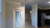 This single-story space showcases a blend of traditional architectural detailing and functional design. The interior is finished with crisp white crown molding that lines the ceilings, adding a polished, classic character to the neutral gray-toned walls. A prominent decorative column with matching molding detail serves as an elegant architectural divider, helping to define separate areas within the open layout while maintaining a sense of visual continuity.

Natural light filters into the space through a white-framed door fitted with horizontal blinds, allowing for adjustable light control throughout the day. The transition from the entryway leads into a carpeted living area, where a ceiling fan with an integrated light fixture provides both comfort and overhead illumination.

The flooring transitions from what appears to be a hard surface near the entry to carpet in the main living area, offering a practical distinction between zones. White baseboards throughout complement the trim work, tying the interior together cohesively. The overall ambiance is clean, bright, and move-in ready, with classic detailing that adds warmth and character to an otherwise neutral palette.