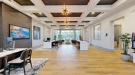 This elegantly designed single-floor space serves as a professional leasing office or community clubhouse, offering a generous and well-appointed interior that balances function with refined aesthetics. The main area is immediately distinguished by striking coffered ceilings with dark wood inlays and eye-catching sputnik-style chandeliers that cast a warm, ambient glow throughout the space. Wide-plank hardwood flooring extends across the entire floor, lending a sense of continuity and warmth.

The reception zone features sleek white workstation desks paired with black chairs, positioned atop patterned area rugs, while a large-format flat-screen television is mounted on a dark wood accent wall nearby. A dedicated lounge area invites visitors to relax in tufted leather armchairs arranged around a round upholstered ottoman, all bathed in natural light pouring through floor-to-ceiling windows that frame outdoor views of a pool and landscaped surroundings.

A beverage station with a built-in coffee machine, white cabinetry, and a compact refrigerator provides added convenience, with a utility room accessible from this area. The entrance foyer features glass double doors and transom windows that maximize natural light. Throughout the space, crisp white walls, curated artwork, and thoughtful lighting create a polished, welcoming atmosphere suited for professional interaction and community engagement.