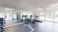 This single-floor fitness facility offers a thoughtfully designed layout divided into two distinct functional zones. The main workout area features durable rubber flooring and is outfitted with a comprehensive selection of equipment, including treadmills, elliptical trainers, stationary cycling bikes, a cable machine, and a dumbbell rack with adjustable bench. Floor-to-ceiling mirrors line the walls, visually expanding the space and supporting proper form during exercise. Recessed panel lighting and ceiling fans maintain a bright, comfortable environment, while large windows along the exterior walls invite generous natural light and provide views of the surrounding outdoor greenery.

An adjoining studio space offers a clean, open area finished with light hardwood-style flooring, ideal for stretching, yoga, or functional training. This room features TRX suspension straps mounted to the wall, recessed skylight panels, and additional large windows that frame outdoor views, creating an airy and motivating atmosphere. A pass-through with interior windows visually connects the studio to the main gym area. Together, the two zones provide a versatile and well-appointed environment suited for a full range of fitness activities.