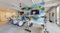 Located at 1401 Aschinger Blvd in Columbus, Ohio, this single-floor fitness center offers a generously sized, thoughtfully designed workout facility. The space is laid out to accommodate a wide range of exercise disciplines, from cardiovascular training to strength conditioning.

The main cardio area showcases a striking, full-wall fitness-themed mural in vivid blues and greens, serving as a dynamic visual backdrop to a row of treadmills, stationary bikes, and elliptical machines, each positioned in front of wall-mounted televisions. Ceiling fans and recessed and track lighting throughout ensure a comfortable and well-lit environment.

The strength training section features a comprehensive selection of cable machines, a free weights rack with dumbbells, weight benches, and plate-loaded equipment. Floor-to-ceiling mirrors line the walls, visually expanding the space and supporting proper form during workouts.

A dedicated functional training area includes a Concept2 rowing machine, resistance bands, plyo boxes, and a multi-functional cable system. This zone is highlighted by a large glass garage-style door that opens directly to an outdoor patio, flooding the space with natural light and fresh air. Large street-facing windows further enhance the bright, open ambiance. Durable, patterned carpet tiles cover the floors throughout, providing a clean, modern aesthetic suited to an active environment.