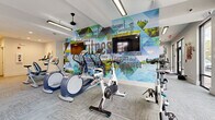 Located at 1401 Aschinger Blvd in Columbus, Ohio, this single-floor fitness center offers a generously sized, thoughtfully designed workout facility. The space is laid out to accommodate a wide range of exercise disciplines, from cardiovascular training to strength conditioning.

The main cardio area showcases a striking, full-wall fitness-themed mural in vivid blues and greens, serving as a dynamic visual backdrop to a row of treadmills, stationary bikes, and elliptical machines, each positioned in front of wall-mounted televisions. Ceiling fans and recessed and track lighting throughout ensure a comfortable and well-lit environment.

The strength training section features a comprehensive selection of cable machines, a free weights rack with dumbbells, weight benches, and plate-loaded equipment. Floor-to-ceiling mirrors line the walls, visually expanding the space and supporting proper form during workouts.

A dedicated functional training area includes a Concept2 rowing machine, resistance bands, plyo boxes, and a multi-functional cable system. This zone is highlighted by a large glass garage-style door that opens directly to an outdoor patio, flooding the space with natural light and fresh air. Large street-facing windows further enhance the bright, open ambiance. Durable, patterned carpet tiles cover the floors throughout, providing a clean, modern aesthetic suited to an active environment.