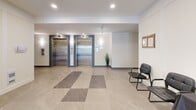Located at 20142 53A Avenue in Langley, BC, this residential building lobby offers a clean, modern aesthetic designed to serve both functional and welcoming purposes. The entrance greets visitors with large-format porcelain tiles laid in a striking checkerboard pattern of light and medium grey tones, creating a polished first impression. Floor-to-ceiling black-framed glass entry doors flood the lobby with natural light, connecting the interior seamlessly to the outdoors.

A dedicated mailroom wall features a built-in array of individual resident mailboxes, neatly recessed into crisp white cabinetry for a streamlined appearance. Adjacent to the entrance, a seating area with chairs and a table provides a convenient space for residents and guests to wait or gather.

Dual stainless steel elevators with blue-grey surround panels serve as the central vertical circulation point, providing efficient access to all residential floors. Directional signage clearly indicates unit ranges, ensuring easy navigation throughout the building. Recessed pot lighting throughout the lobby creates a bright, inviting atmosphere without harsh glare.

The hallway extending from the lobby is finished with neutral-toned carpet and continuous wall sconces, maintaining a consistent and welcoming ambiance down its length. The overall design is contemporary and functional, offering residents a well-maintained and accessible common space.