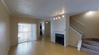 This two-story residence offers a thoughtfully designed layout that balances comfort and functionality across both levels. The main living area serves as the heart of the home, showcasing smooth hardwood-style flooring that extends throughout the open space. A classic fireplace with a clean white mantel anchors one wall, providing a welcoming focal point for the room. Track lighting overhead ensures the space is well-lit, complementing the natural light that pours in through large sliding glass doors dressed with vertical blinds. The textured ceiling adds a traditional architectural detail found throughout the main floor. A staircase with a white railing leads to the upper level, offering a seamless transition between floors. Adjacent to the living area, a ceiling fan is visible in what appears to be a dining or secondary living space, hinting at the home's open-concept flow. The kitchen can be glimpsed in the background, featuring dark cabinetry that provides contrast to the light-toned walls and flooring. Overall, the space presents a neutral, move-in-ready palette with practical finishes and a layout well-suited for everyday living.