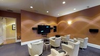 Located at 3800 Bostic Dr in Greenville, NC, this single-floor space showcases a thoughtfully designed media lounge that blends comfort with style. The room is defined by its rich, warm-toned walls adorned with sweeping, wave-like artistic murals in golden hues, creating an inviting and visually dynamic ambiance. Recessed ceiling lighting provides soft, even illumination throughout, complemented by a wall-mounted accent light that adds a warm glow to the space. The centerpiece of the room is a large wall-mounted flat-screen television flanked by surround sound speakers, making it well-suited for an immersive viewing experience. Surrounding the screen, built-in media storage adds a practical touch. Seating consists of a collection of cushioned swivel chairs upholstered in a neutral, patterned fabric, arranged to face the screen and promote a communal atmosphere. Each chair is paired with a small round side table, offering convenience for guests. The dark carpeted floor grounds the warm color palette and contributes to the room's cozy, theater-like feel. Overall, this media room offers a refined and comfortable environment ideal for entertainment and social gatherings.