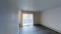 This single-floor space is designed with a straightforward, functional layout. The main living area is a standout feature, offering a generous, open feel enhanced by neutral white walls and smooth ceilings. Dark-toned, hardwood-style flooring runs throughout, providing a modern contrast to the light-colored walls and adding a sense of warmth and continuity to the space.

A large sliding glass door dominates one wall of the living area, fitted with vertical blinds that allow for adjustable light control. The door opens to a balcony, offering views of surrounding trees and greenery, bringing natural light into the interior. A wall-mounted air conditioning unit sits conveniently above the sliding door, while baseboard heaters line the walls, ensuring year-round climate comfort.

An open doorway connects the living area to an adjacent room, suggesting a flexible, flowing layout between spaces. The overall ambiance of the space is clean and neutral, providing a blank canvas that accommodates a variety of furnishing styles and personal aesthetics. The combination of natural light, outdoor access, and modern flooring gives the space a bright, practical character.
