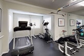 This dedicated fitness space is thoughtfully designed to accommodate a full range of exercise activities within a single floor. The room features dark rubber flooring throughout, providing both durability and traction during workouts. A large wall-to-wall mirror installation spans one side of the space, visually expanding the room while allowing users to monitor their form during exercise.

The cardio section is equipped with a treadmill, elliptical trainer, and stationary bike, positioned near multiple windows that allow natural light to filter into the space. A multi-station cable machine serves as the centerpiece for strength training, complemented by a weight bench and additional resistance equipment. A medicine ball rack stocked with various weighted balls adds versatility for functional training exercises.

Crown molding along the ceiling adds a refined architectural touch, while a textured ceiling houses flush fluorescent light panels that ensure consistent illumination throughout. Wall-mounted flat-screen televisions are positioned for visibility from multiple workout stations. Framed artwork decorates the neutral gray walls, contributing to a clean, motivating atmosphere. A dedicated entry door provides direct access to the space, and a fire extinguisher is conveniently mounted near the exit for safety.