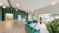 Located at 1850 Paisley Place in New Castle, Delaware, this single-floor community space makes a striking first impression with its dramatic vaulted ceilings and an eye-catching deep green accent wall adorned with a bold geometric chevron pattern in white. The expansive open-plan living room is finished with light-toned hardwood flooring that runs throughout, creating a clean, contemporary aesthetic. Oversized windows along multiple walls flood the interior with abundant natural light and offer views of the surrounding neighborhood. Stylish globe pendant lights hang at varying heights, adding a warm, modern touch to the lofty ceiling. The lounge area is anchored by teal leather sofas and plush green accent chairs arranged around a curved area rug, providing an inviting gathering space. Sage green painted walls in the central zone complement the bolder accent wall, contributing to a cohesive, nature-inspired color palette. Interior pass-through windows connect the main living area to an adjacent fitness room, which is visible and accessible from the lounge, equipped with cardio machines. Lush potted tropical plants are thoughtfully placed throughout, enhancing the fresh, vibrant atmosphere of this well-appointed community amenity space.