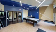 The Spectrum Apartments Game Room is a lively and well-appointed recreational amenity designed to bring residents together for leisure and entertainment. The space is entered through a set of glass-paneled doors, with a branded welcome mat and a vending machine conveniently positioned at the entry. Inside, bold blue and warm tan accent walls create an energetic, modern atmosphere, complemented by striking geometric wall designs and contemporary linear pendant lighting overhead. The main game area centers around a full-size pool table and a shuffleboard table, providing ample space for competitive play on durable hardwood-style flooring. Adjacent to the game zone, a kitchenette counter with a stainless steel top and built-in sink offers a functional refreshment station, while a wall-mounted flat-screen TV adds an entertainment focal point. Colorful, curved lounge seating and decorative wall art add personality and comfort to the space. Recessed lighting supplements the overhead fixtures, ensuring the entire room is well-lit throughout. The open layout flows seamlessly between zones, making this game room an inviting destination for socializing, recreation, and relaxation within the Spectrum Apartments community.