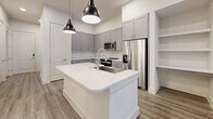 Located at 10400 Research Blvd in Austin, TX, this thoughtfully designed unit offers a compact yet functional single-floor layout. The open-concept kitchen and living area serves as the heart of the space, featuring warm wood-look flooring, crisp white walls, and recessed lighting throughout. The kitchen is a standout, appointed with gray shaker-style cabinetry, quartz countertops, a large stainless steel farmhouse sink set in a central island, sleek pendant lighting, a classic white tile backsplash, and a full suite of stainless steel appliances including a refrigerator, range, microwave, and dishwasher. Open shelving provides additional storage. The living area is bright and airy, with natural light streaming through French doors that lead directly to a private patio. The patio features artificial turf and metal railing, with views of the building's shared courtyard, which includes a brick fireplace, pergola-covered seating, and communal outdoor furnishings. A dedicated in-unit laundry room houses a full-size front-loading washer and dryer with overhead shelving. The exterior of the building showcases a modern mix of brick and dark lap siding, lending a polished, contemporary aesthetic to this Austin residence.