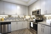 This well-designed single-floor residence offers a cohesive blend of modern finishes and functional living spaces. The kitchen is a standout feature, equipped with sleek flat-panel cabinetry in a warm grey tone, a mosaic tile backsplash, light quartz countertops, and a full suite of stainless steel appliances including a refrigerator with ice maker, range, microwave, and dishwasher. A generously sized center island with pendant lighting provides additional prep space and casual seating, making it ideal for both cooking and entertaining.

The open living area is bathed in natural light from large corner windows dressed with Roman shades, offering views of the surrounding exterior. A ceiling fan, hardwood-style flooring, and a welcoming layout with ample seating space define this bright and airy room, which also provides access to an outdoor balcony.

The two bedrooms are thoughtfully designed, each featuring plush carpeting, natural light from windows with blinds, and ceiling fans for comfort. One bedroom features a bold navy accent wall, while the other maintains a softer grey palette, both lending distinct character to their spaces.

The bathrooms are finished with clean, contemporary materials — one offering a soaking tub with full tile surround, and another featuring a light wood vanity with a white countertop and a large framed mirror.