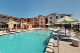 Located at 148 Peabody Rd in Vacaville, CA, this outdoor amenity area is a well-appointed recreational space designed for relaxation and leisure. The centerpiece is a generously sized swimming pool with crystal-clear turquoise water, complemented by a separate hot tub/spa for added comfort. The expansive concrete pool deck is lined with cushioned lounge chairs and shaded by multiple large market umbrellas, providing ample space for sunbathing under the abundant California sunshine. A covered pergola structure offers additional shade and houses an outdoor BBQ grill station, making it ideal for outdoor gatherings and casual dining. The area is fully enclosed by black metal perimeter fencing, ensuring privacy and security. Tasteful landscaping with potted plants and greenery adds a welcoming touch throughout. The surrounding multi-story residential building frames the space, with balconies overlooking the pool area. A dedicated bathroom facility serves the outdoor space. The clean, modern design and well-maintained surfaces reflect a commitment to quality, while the open layout and abundant natural light create an inviting atmosphere perfect for year-round outdoor enjoyment in the warm Vacaville climate.