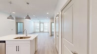Located at 17501 Dallas Parkway in Dallas, TX, this well-appointed two-bedroom, two-bathroom residence offers a bright and modern living experience across a single floor. The open-concept kitchen and living area serves as the heart of the home, showcasing white shaker cabinetry, a textured subway tile backsplash, stainless steel appliances, and a large center island with a single-basin sink and pendant lighting. Light wood-style flooring flows throughout the main living spaces, lending warmth and continuity to the design. The living area is flooded with natural light through large windows flanking a glass-panel door, fitted with white horizontal blinds. Both bedrooms are carpeted and include ceiling fans, generous windows, and dedicated walk-in closets with built-in shelving and hanging rods for ample storage. The primary bathroom features a dual-sink vanity with white cabinetry, black hardware accents, light countertops, an illuminated mirror, and wood-style flooring, with direct access to the primary bedroom. A second full bathroom and an additional closet round out the layout. Clean white walls and recessed lighting throughout contribute to a bright, contemporary ambiance well-suited for comfortable Dallas living.