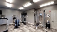 This fitness room offers a functional and well-organized workout space designed to support a variety of exercise routines. The room is finished with neutral gray walls and complementary patterned carpet tile flooring, creating a clean, modern aesthetic throughout. Bright flush-mount fluorescent panel lighting illuminates the space evenly from the ceiling, ensuring excellent visibility during workouts.

The room is equipped with a selection of cardio machines, including an elliptical trainer and a stationary recumbent bike, both from Life Fitness — a reputable commercial-grade brand. A multi-function cable resistance machine anchors one side of the room, accompanied by an adjustable weight bench, making it suitable for a range of strength training exercises. The cable system also includes an integrated display screen for guided workouts.

A wall-mounted flat-screen television is positioned for easy viewing from any point in the room, adding an entertainment option during exercise sessions. A small window provides a modest source of natural light. The overall layout is efficient and thoughtfully arranged, maximizing usability within the available footprint.