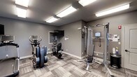 This fitness room offers a functional and well-organized workout space designed to support a variety of exercise routines. The room is finished with neutral gray walls and complementary patterned carpet tile flooring, creating a clean, modern aesthetic throughout. Bright flush-mount fluorescent panel lighting illuminates the space evenly from the ceiling, ensuring excellent visibility during workouts.

The room is equipped with a selection of cardio machines, including an elliptical trainer and a stationary recumbent bike, both from Life Fitness — a reputable commercial-grade brand. A multi-function cable resistance machine anchors one side of the room, accompanied by an adjustable weight bench, making it suitable for a range of strength training exercises. The cable system also includes an integrated display screen for guided workouts.

A wall-mounted flat-screen television is positioned for easy viewing from any point in the room, adding an entertainment option during exercise sessions. A small window provides a modest source of natural light. The overall layout is efficient and thoughtfully arranged, maximizing usability within the available footprint.