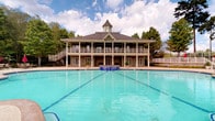 Located at 5001 River Chase Dr in Phenix City, Alabama, this community clubhouse is a standout centerpiece defined by its classic Southern architectural character. The building rises with a distinctive cupola at its peak, a steeply pitched dark roof, and wide wrap-around covered balconies supported by decorative columns on both levels, evoking a timeless, plantation-inspired aesthetic. Multiple sets of French doors line the ground level, opening to the expansive pool deck and inviting seamless indoor-outdoor flow. A generous outdoor gathering area sits just outside the building's entrance, complete with a fireplace or grill feature, potted plantings, and comfortable seating — ideal for social events and relaxation. The centerpiece of the outdoor space is a large, resort-style swimming pool with clearly marked lap lanes, complemented by an elevated spa or fountain feature finished in rich cobalt blue tile. The pool deck is finished in warm stone pavers, and the entire area is framed by mature trees that provide natural privacy and shade. A staircase on the exterior connects the ground level to the upper balcony, offering elevated views of the pool and surrounding landscape. The overall ambiance is welcoming, refined, and well-suited for an active community lifestyle.