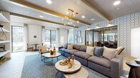 This single-floor amenity space is designed with a cohesive, contemporary aesthetic that balances comfort and productivity. The main lounge area serves as the centerpiece, featuring a generously sized sectional sofa, layered with accent pillows, set atop a blue patterned area rug over light wood-style flooring. An eye-catching brass sputnik chandelier hangs from a ceiling accented with exposed beams, adding a modern yet warm character to the space. Large floor-to-ceiling windows invite abundant natural light and offer views of the surrounding exterior grounds. A textured tile accent wall with glass-panel partitions separates the lounge from an adjoining conference or co-working nook, maintaining visual openness while defining distinct zones. The library-style workspace features an entire wall of built-in open shelving styled with books, plants, and décor, flanked by individual desk stations with task lighting and ergonomic seating. A separate business center room offers a streamlined countertop workspace with teal cabinetry, quartz surfaces, and curated wall art, ideal for focused individual work. A dining-height round table with mid-century modern chairs sits near a large window grouping, creating a bright and versatile meeting area. Throughout, the design palette of soft neutrals, teal accents, and warm gold fixtures creates an inviting and polished communal environment.