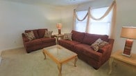 This single-floor space centers around a welcoming living room that exudes a warm, relaxed ambiance. The room is furnished with two generously sized sofas upholstered in a rich brown fabric, accented with decorative patterned throw pillows that add visual interest. A glass-top coffee table with an ornate wooden base serves as the focal point of the seating arrangement, offering both style and functionality. A matching end table holds a warm-toned table lamp, contributing to the room's cozy atmosphere. Light-colored carpeting runs throughout, softening the overall feel of the space. Natural light filters in through a window dressed with elegant sheer draping, adding an airy quality to the room. The walls are painted in a soft, neutral tone that complements the warm furnishings and creates a cohesive, inviting environment. The layout is thoughtfully arranged to encourage conversation and comfort, making it well-suited for everyday living or casual entertaining.