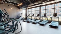 Located at 301 Winchester Ave in New Haven, CT, this contemporary fitness center offers a thoughtfully designed exercise space across a single floor. The facility is divided into distinct functional zones, each tailored to a different aspect of physical training.

The main cardio area features a generous row of treadmills and elliptical machines positioned along a wall of large, floor-to-ceiling windows that flood the space with natural light and provide views of the surrounding neighborhood. The exposed ceiling reveals industrial-style ductwork and conduit, complemented by sleek linear pendant lighting that reinforces the space's modern, urban aesthetic. Durable speckled rubber flooring throughout the cardio zone provides both safety and resilience.

An adjoining open studio area showcases wide-plank hardwood flooring and dark accent walls lined with large mirrors, creating a bright and expansive atmosphere ideal for stretching, yoga, or group workouts. This zone also houses a functional training rig equipped with a punching bag, medicine balls, kettlebells, resistance bands, and stability equipment, all organized for easy access.

Together, the zones create a comprehensive, well-appointed fitness facility that balances high-performance equipment with a polished, industrial-modern design sensibility.