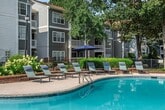 This single-floor, 3-bedroom, 2-bathroom apartment offers 1,020 square feet of well-organized living space. The open living room is finished with light hardwood-style flooring, neutral beige walls, and a textured ceiling. A sliding glass door leads to a private balcony overlooking a landscaped courtyard with mature trees and manicured shrubs, allowing natural light to fill the room. The kitchen features a central island with a marble-look countertop, stainless steel appliances including a refrigerator and dishwasher, and a built-in sink — all set over matching hardwood-style flooring. The three bedrooms are carpeted in neutral tones, with large windows that bring in natural light and offer outdoor views. The primary bedroom measures 150 square feet and includes closet access. Both bathrooms are identically appointed with white tiled tub-shower combinations, white vanity cabinetry, and hardwood-style flooring, providing a clean and consistent aesthetic. A dedicated laundry room houses a full-size washer and dryer with overhead wire shelving for additional storage. Three closets throughout the unit provide ample storage. The overall ambiance is clean, neutral, and functional.