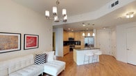 This single-floor space showcases a thoughtfully designed open-concept layout that seamlessly connects the living and kitchen areas, creating a cohesive and functional environment. The living area is anchored by a contemporary multi-arm chandelier that adds a refined focal point to the space, complementing the smooth, light-toned ceilings accented with recessed lighting. Warm hardwood-style flooring flows throughout, tying the areas together with a clean, modern aesthetic.

The kitchen is outfitted with light maple cabinetry, dark granite countertops, and a full suite of stainless steel appliances, including a French door refrigerator, range, and built-in microwave. A dark tile backsplash adds contrast and visual interest against the cabinetry. Pendant lights hang above a central kitchen island topped with granite, which features an integrated sink and is paired with sleek white bar stools, making it ideal for casual dining or entertaining.

The open flow between the kitchen and living space allows for easy interaction and maximizes the sense of openness throughout. Recessed lighting in the kitchen ensures the workspace is well-lit, while the overall design balances practicality with a contemporary, welcoming ambiance suited for everyday living.