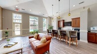 Located at 1660 Hampton Knoll Drive in Akron, Ohio, this single-floor space combines a living room and kitchen into one cohesive, thoughtfully designed area. The living room is anchored by a tile-surround fireplace flanked by built-in shelving, creating a warm and inviting focal point. Vaulted ceilings rise above, adding a sense of openness and grandeur. Neutral taupe and gray tones coat the walls, complemented by classic white chair-rail wainscoting that runs throughout, lending a refined, traditional character to the space. Hardwood-style plank flooring flows seamlessly from the living area into the kitchen, tying the two zones together. Large windows allow natural light to pour in, brightening the entire floor plan. The kitchen is well-appointed with rich medium-toned wood cabinetry, dark granite-style countertops, and a central island with bar seating for multiple guests. Pendant lantern-style light fixtures hang from the soaring ceiling above the kitchen, adding warmth and a craftsman-inspired aesthetic. Ample counter space and additional lower cabinetry provide generous storage and functionality. Together, the living and kitchen areas form a comfortable, versatile space ideal for gathering and everyday use.