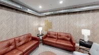 This single-floor space is thoughtfully designed to serve as a functional communal area, combining a lounge and a full kitchen into one cohesive layout. The lounge area is furnished with rich cognac-toned leather sofas and accent chairs arranged around small side tables, creating an inviting setting for conversation and relaxation. The walls are dressed in a textured chevron-patterned wallcovering in warm neutral tones, adding visual interest to the space. Exposed silver HVAC ductwork runs along the upper perimeter, lending an industrial contrast to the otherwise warm interior. Recessed lighting and floor lamps provide layered illumination throughout. Adjacent to the lounge, a dining area with wooden chairs and small tables offers additional seating. The kitchen is well-appointed with dark espresso shaker-style cabinetry, white countertops, and a full suite of stainless steel appliances, including a refrigerator, electric range with a range hood, microwave, dishwasher, and a double-basin sink. Light wood-tone flooring anchors the kitchen zone, while patterned carpet defines the lounge. Together, these areas create a practical and comfortable environment suited for shared everyday use.