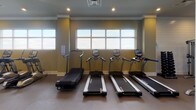 This fitness center is thoughtfully designed to accommodate a wide range of workout styles and fitness goals. The space is organized into distinct zones, each serving a specific purpose while maintaining a cohesive, modern aesthetic throughout. Walls are finished in a warm olive-green tone complemented by crisp white shiplap accents, and recessed lighting provides consistent illumination across the entire floor.

The cardio zone lines one wall beneath a row of wide, multi-pane windows that allow ample natural light to fill the space. This area houses a lineup of treadmills, elliptical trainers, and a rowing machine, all positioned to take advantage of the outdoor views. Adjacent to the cardio section, a second windowed alcove offers additional cardio equipment, including ellipticals and a stair climber.

At the heart of the facility stands a multi-functional training rig equipped with battle ropes, suspension trainers, and a tiered rack stocked with medicine balls and kettlebells. A ceiling fan overhead keeps air circulating during high-intensity sessions. A water fountain is conveniently mounted nearby, and a wall-mounted flat-screen television adds an entertainment option.

A glass-walled studio room is visible from the main floor, offering a separate environment for group classes or individual training, with mirrors and additional equipment inside. The durable rubber flooring throughout ensures safety and comfort, making this fitness center a comprehensive and well-appointed facility.