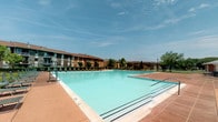 This outdoor amenity space centers around a generously sized swimming pool with clear, turquoise water, designed to serve a multi-building residential community. The pool area is finished with a broad, tiled concrete deck in warm terracotta tones, providing ample space for lounge seating and recreational use along the pool's perimeter. Rows of green lounge chairs line one side of the deck, offering dedicated relaxation areas for residents enjoying sunny days outdoors.

The surrounding landscape features mature trees that provide natural greenery and partial shade, complementing the open-air setting. The pool is enclosed by a perimeter fence, ensuring safety and privacy. Brick construction is prominently featured throughout the exterior elements of the adjacent buildings and facility structures, contributing a classic, durable aesthetic to the overall environment.

The multi-story residential buildings visible in the background are constructed with brick facades and feature pitched rooflines, creating a cohesive architectural character across the community. Entry points to the pool area are accessed through gated passages framed by brick walls and painted paneled doors. The space benefits from abundant natural light and open sky views, making it a standout communal outdoor feature well-suited for warm-weather enjoyment.