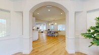 This well-appointed single-floor space offers a thoughtfully designed layout that balances functionality with a warm coastal aesthetic. The entry area welcomes visitors with soaring ceilings, large windows that invite abundant natural light, and a clean, airy atmosphere complemented by light hardwood flooring that extends throughout the entire space.

A graceful arched passageway connects the entry to the main areas, which include an open lounge and a dedicated meeting or workspace. The lounge features a textured white brick accent wall, recessed lighting, and a relaxed seating arrangement anchored by woven rattan chairs and a crisp white coffee table. Double glass-paned doors lead directly to a fitness room equipped with exercise equipment, visible from the main lounge area.

The meeting area is outfitted with a substantial light-wood conference table, woven pendant lighting, and ample natural light from multiple windows, making it ideal for collaborative work.

The kitchenette stands out with its striking powder-blue glazed tile backsplash, white shaker cabinetry with gold hardware, white quartz countertops, and stainless steel appliances. Throughout, the design palette of crisp white walls, soft neutrals, and natural wood tones creates a cohesive, calming environment suited for a professional or community-oriented setting.
