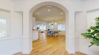 This well-appointed single-floor space offers a thoughtfully designed layout that balances functionality with a warm coastal aesthetic. The entry area welcomes visitors with soaring ceilings, large windows that invite abundant natural light, and a clean, airy atmosphere complemented by light hardwood flooring that extends throughout the entire space.

A graceful arched passageway connects the entry to the main areas, which include an open lounge and a dedicated meeting or workspace. The lounge features a textured white brick accent wall, recessed lighting, and a relaxed seating arrangement anchored by woven rattan chairs and a crisp white coffee table. Double glass-paned doors lead directly to a fitness room equipped with exercise equipment, visible from the main lounge area.

The meeting area is outfitted with a substantial light-wood conference table, woven pendant lighting, and ample natural light from multiple windows, making it ideal for collaborative work.

The kitchenette stands out with its striking powder-blue glazed tile backsplash, white shaker cabinetry with gold hardware, white quartz countertops, and stainless steel appliances. Throughout, the design palette of crisp white walls, soft neutrals, and natural wood tones creates a cohesive, calming environment suited for a professional or community-oriented setting.