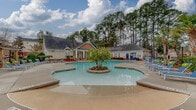 Located at 1450 Bluewater Way in Charleston, SC, this community amenity space at Colton's Landing Apartments centers around a generously sized freeform swimming pool with a distinctive landscaped island feature at its center. The pool area is designed for both relaxation and socializing, with an expansive concrete deck surrounding the water and ample lounge seating, including colorful chaise lounges arranged throughout the space. A standout stone outdoor fireplace anchors one end of the pool area, providing a cozy gathering spot framed by tall pines and palm trees that create a natural, private backdrop. The single-story clubhouse building is finished in classic Charleston-style yellow siding with a charcoal roof, dormer accents, and a welcoming covered porch supported by elegant white columns set on stacked stone bases. The porch is furnished with comfortable all-weather seating and offers direct views of the pool. French doors lead into the interior of the clubhouse, and a separate auxiliary structure provides additional poolside facilities. A pergola-shaded seating area extends along the deck, enhancing the resort-like atmosphere. The entire space is enveloped by mature pine and palm trees, lending a lush, wooded ambiance to this outdoor community retreat.
