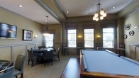 This single-floor recreation and game room offers a refined yet welcoming atmosphere designed for social gatherings and leisure activities. The space is anchored by a full-sized billiards table finished in rich wood tones with a slate-green felt surface, positioned prominently on hardwood flooring. Adjacent to it, a carpeted seating area accommodates a round game table surrounded by upholstered chairs, ideal for card games or casual meetings.

The room features elevated ceilings with decorative coffered detailing, enhancing the sense of openness and architectural character. Warm-toned walls are accented by classic wainscoting panels, contributing to the space's traditional, club-like aesthetic. Illumination is thoughtfully layered, combining elegant multi-bulb pendant chandeliers, recessed ceiling lights, and decorative wall sconces that collectively create an inviting ambiance.

Large double-hung windows dressed with fabric valances line the perimeter walls, allowing generous natural light to fill the space and offering views of the surrounding exterior landscape. Bar-height stools positioned near the windows provide additional casual seating. Overall, the room balances functionality with a polished, upscale design sensibility.