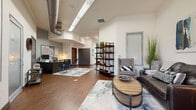 This single-floor space combines industrial and contemporary design elements to create a welcoming, functional environment. Soaring ceilings with exposed silver ductwork lend an urban, loft-inspired character throughout, while rich hardwood-style flooring anchors each zone with warmth and cohesion.

The main entry area serves a dual purpose, featuring a dedicated workstation with a desktop computer and seating for consultations, as well as a wall-mounted property map for reference. This workspace flows seamlessly into a comfortable lounge area, where an open-back wood-and-metal shelving unit acts as a natural divider. The lounge is furnished with a leather loveseat, upholstered accent chairs, and a round wood-top coffee table set atop a layered area rug, creating an inviting gathering space. Abstract artwork in blues and teals adds a curated, artistic touch to the walls.

A second, more intimate sitting area features a bold slate-gray accent wall with a wall-mounted flat-screen television, a wood media console, and a mix of upholstered sofas and leather chairs arranged around a concrete-top coffee table — ideal for informal meetings or relaxation.

The kitchenette is appointed with two-tone cabinetry in gray and espresso, granite countertops, a stainless steel refrigerator, microwave, and coffee station, offering convenient amenities for staff and visitors alike.