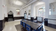 This distinguished single-floor space showcases a seamless blend of classical architecture and contemporary design. Soaring ceilings adorned with intricate plaster cornices and ornamental columns establish a stately atmosphere throughout. Dramatic crystal chandeliers provide warm, elegant illumination, complementing the natural light that pours in through large arched and multi-pane windows, offering views of lush greenery beyond.

The main gathering area features a substantial dark wood conference table surrounded by plush navy blue and gold-framed chairs, making it well-suited for meetings or collaborative work. Glossy terrazzo flooring flows through the corridors and entry areas, while dark hardwood anchors the central room, adding visual contrast and warmth.

Bold dark wainscoting lines the walls throughout, paired with crisp white upper walls that heighten the sense of vertical space. Arched doorways and Gothic-influenced architectural details lend a sense of heritage and character. Built-in mailbox units and French doors with decorative crests provide functional and aesthetic focal points near the entrance. A graceful wrought iron stair railing adds an artisan touch. The overall ambiance is refined, professional, and architecturally impressive.