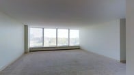 This single-floor space is defined by its expansive wall of floor-to-ceiling windows that stretch across the entire width of the main living area, flooding the interior with abundant natural light. The panoramic windows frame elevated views of a lush tree canopy and surrounding urban skyline, creating a strong visual connection to the outdoors. The room is generously proportioned, offering a wide, open layout well-suited for flexible furniture arrangements. Neutral-toned wall-to-wall carpeting covers the floor throughout, contributing to a clean and understated aesthetic. The crisp white walls and smooth ceiling enhance the sense of brightness and airiness. A continuous baseboard heating unit runs along the base of the window wall, a practical feature that ensures even warmth throughout the colder months. The overall ambiance is calm and uncluttered, with the abundance of natural light and elevated outdoor vistas serving as the defining characteristics of the space. The minimalist finishes and open floor plan provide a versatile canvas ready to accommodate a variety of interior design approaches.