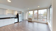 This contemporary single-floor space showcases a thoughtfully designed open-concept layout that seamlessly integrates the living and kitchen areas. Large, floor-to-ceiling windows dominate the exterior wall, flooding the interior with an abundance of natural light and offering clear views of the surrounding urban streetscape. The clean white walls and smooth ceilings complement the warm, wood-style flooring that extends throughout the main living area, creating a cohesive and inviting atmosphere.

The kitchen is efficiently arranged along one wall and features light-toned cabinetry, a stainless steel refrigerator, a dishwasher, and dark countertops that provide a modern contrast to the bright cabinetry above. A dedicated entry corridor leads to the main living space, adding a sense of defined transition between areas.

A wall-mounted climate control unit is positioned beneath the windows, ensuring year-round comfort. Recessed ceiling lighting supplements the natural light, maintaining a well-lit environment throughout the day and evening. The overall design aesthetic is clean, modern, and minimalist, making the space feel open and functional. The urban views visible through the expansive windows further enhance the connected, contemporary character of this well-appointed space.