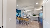 This single-floor residence offers a thoughtfully designed open-concept layout that seamlessly connects the kitchen, dining, and living areas. The kitchen is a standout feature, showcasing grey flat-panel cabinetry with matte black hardware, crisp white quartz countertops, and a textured white tile backsplash. Stainless steel appliances, including a range, microwave, and refrigerator, complement the space, while a central island with pendant lighting and woven rattan counter stools provides casual dining. Recessed lighting and subtle cove lighting above the cabinetry add warmth throughout. Light wood-style flooring runs through the main living areas, contributing to a clean, contemporary aesthetic.

The living room makes a bold impression with a deep teal accent wall, natural light from nearby windows, and ample space for comfortable seating. The bedroom is carpeted and generously sized, featuring a large window with outdoor views. Bathrooms are finished with white quartz vanities, grey cabinetry, matte black fixtures, and wood-style flooring. A dedicated laundry area houses a full-size washer and dryer with overhead shelving, adding practical convenience to this well-appointed residence.