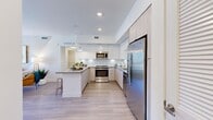 This well-appointed single-story residence in Port Saint Lucie, Florida, offers a thoughtfully designed open floor plan with modern finishes throughout. The living area is bright and welcoming, featuring light wood-style flooring, a ceiling fan, and a large sliding glass door that opens to a private balcony with views of the surrounding community. An accent wall with geometric tonal detailing adds a distinctive architectural touch to the living space.

The kitchen is a standout feature, showcasing sleek two-tone cabinetry — combining glossy white upper cabinets with light wood lower cabinets — paired with a classic white subway tile backsplash, quartz countertops, and a full suite of stainless steel appliances including a refrigerator, range, microwave, and dishwasher. A central island with bar seating provides additional prep space and casual dining options. Recessed lighting and pendant fixtures illuminate the space effectively.

The bedrooms are comfortably sized with light wood-style flooring, neutral walls, and natural light from well-placed windows. The bathroom is clean and contemporary, featuring a single-sink vanity with light wood cabinetry, a large frameless mirror, and a tiled shower-tub combination. Throughout the residence, the design reflects a cohesive modern aesthetic suited for comfortable Florida living.