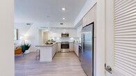 This well-appointed single-story residence in Port Saint Lucie, Florida, offers a thoughtfully designed open floor plan with modern finishes throughout. The living area is bright and welcoming, featuring light wood-style flooring, a ceiling fan, and a large sliding glass door that opens to a private balcony with views of the surrounding community. An accent wall with geometric tonal detailing adds a distinctive architectural touch to the living space.

The kitchen is a standout feature, showcasing sleek two-tone cabinetry — combining glossy white upper cabinets with light wood lower cabinets — paired with a classic white subway tile backsplash, quartz countertops, and a full suite of stainless steel appliances including a refrigerator, range, microwave, and dishwasher. A central island with bar seating provides additional prep space and casual dining options. Recessed lighting and pendant fixtures illuminate the space effectively.

The bedrooms are comfortably sized with light wood-style flooring, neutral walls, and natural light from well-placed windows. The bathroom is clean and contemporary, featuring a single-sink vanity with light wood cabinetry, a large frameless mirror, and a tiled shower-tub combination. Throughout the residence, the design reflects a cohesive modern aesthetic suited for comfortable Florida living.